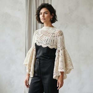 May include: A cream-colored crochet bolero featuring an intricate fan-like stitch pattern, a high neckline, and wide, flared bell sleeves. The garment is worn over a black sleeveless top and black trousers, showcasing a delicate, openwork texture.