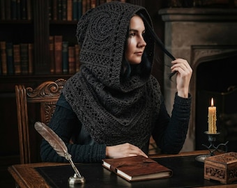 May include: A dark charcoal grey crochet hooded cowl featuring an intricate textured pattern and a prominent crescent moon motif integrated into the hood. The garment is crafted with a thick yarn, providing a structured drape around the head and shoulders.