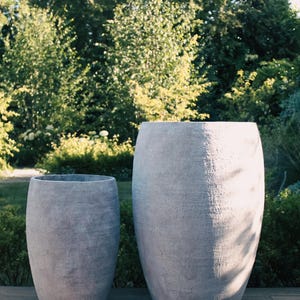 May include: Two large, grey, textured planters with a weathered appearance. The planters are cylindrical with a slightly tapered shape. The background features lush green foliage and a wooden deck.