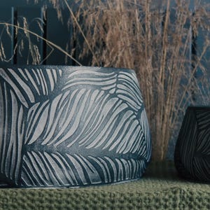 May include: Two dark grey ceramic planters with a textured, carved leaf design. The larger planter is oval-shaped, and the smaller one is rectangular. Dried ornamental grass is in the background, with a teal wall.