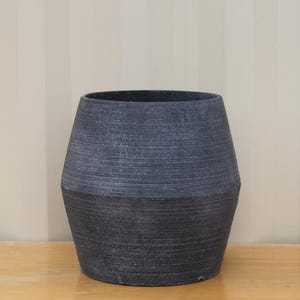 May include: A dark charcoal grey planter with a wide, rounded body that tapers towards the base and the top. The exterior surface features a distinct horizontal ribbed texture throughout, creating a matte finish with subtle variations in colour.