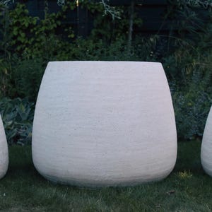 May include: Three off-white, rounded planters with a textured, matte finish. The planters have a wide, open top and a tapered base, arranged in a row with the largest one positioned in the centre, showcasing a consistent, rough surface texture.