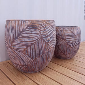 May include: Two rounded planters featuring a textured surface with a repeating embossed leaf pattern. The finish is a weathered copper-brown tone with dark grey recessed areas, creating a distinct contrast across the curved exterior of each pot.