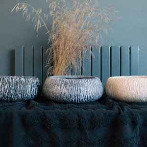 May include: Three textured ceramic bowls in shades of black, white, and beige. The bowls are displayed on a dark green cloth, with dried ornamental grass in the background. The bowls are suitable for home decor.