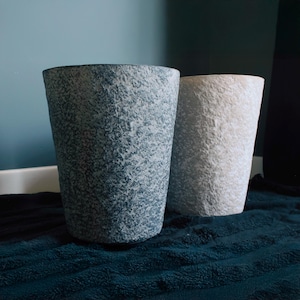 May include: Two cylindrical planters with a rough, stone-like texture. One planter is a dark charcoal grey, while the other is a light off-white. Both vessels feature a tapered shape that narrows towards the base, showcasing a rugged exterior finish.