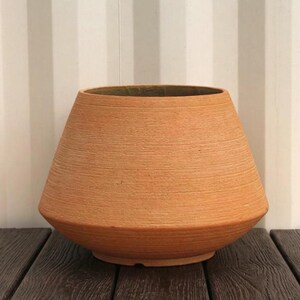 May include: A wide, tapered terracotta planter with a textured, horizontally ribbed surface. The pot features a broad, open top and a narrower base, showcasing the natural, earthy orange-brown colour of the clay material throughout its exterior.