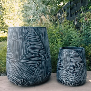May include: Two dark grey planters with a leaf pattern. The larger planter is approximately 61 cm tall and 46 cm wide, while the smaller one is about 46 cm tall and 36 cm wide. Both are on a wooden deck, with greenery in the background.