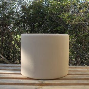 May include: A cylindrical planter with a smooth, matte, light beige finish. The vessel features a simple, clean design with straight vertical walls and a flat, circular top opening, presenting a uniform and neutral appearance throughout.