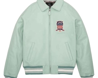 May include: A light green leather jacket with a collared neckline and a zipper closure. The jacket features a patch on the chest with the text "AVIREX" and an American flag design. The cuffs and hem have a striped design.