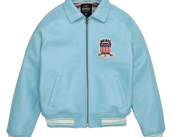 May include: Light blue leather jacket with a zip-up front, collar, and ribbed cuffs and hem. Features a patch on the chest with the text "AVIREX" and a patriotic design. The jacket has two front pockets.