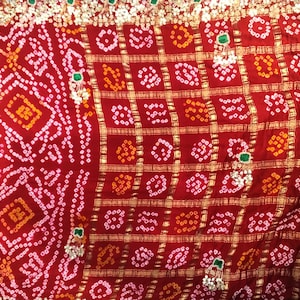 May include: A detailed red textile with a complex pattern. The design incorporates geometric shapes and dotted patterns, highlighted with gold detailing. Small green and white decorative elements are also present. The fabric has a rich, textured appearance.
