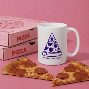 Könnte beinhalten: Ein weißer Keramikbecher mit einer violetten Grafik eines Pizzastücks, das von einem Auge in einem Dreieck gekrönt wird, darunter der Text "IN CRUST WE TRUST". Der Becher steht neben zwei Pizzastücken und zwei gestapelten rosa Pizzakartons.