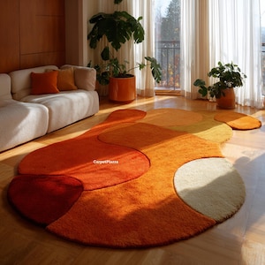 Op de afbeelding: Een abstract vloerkleed in tinten oranje, rood en crème. Het kleed heeft een unieke, organische vorm met meerdere overlappende secties. De zachte textuur en warme kleuren creëren een gezellige en uitnodigende sfeer in een woonkamer.
