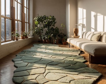 May include: A large, green, stone-patterned rug on a hardwood floor. The rug has varying shades of green and a unique, organic shape. The room features large windows, plants, and a neutral-toned sofa.