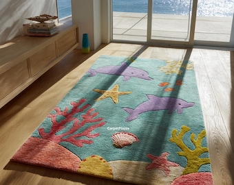 Op de afbeelding: Een rechthoekig vloerkleed met een aquatisch thema. Het kleed heeft een lichtblauwe achtergrond met koraal, zeesterren en twee paarse dolfijnen. Een zeeschelp en vissen zijn ook afgebeeld. Het kleed ligt op een houten vloer.