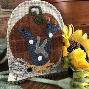 May include: A brown and white felt pumpkin with a blue and green bird design. The pumpkin has a bell on top and a star at the bottom. The pumpkin is sitting on a wooden surface.