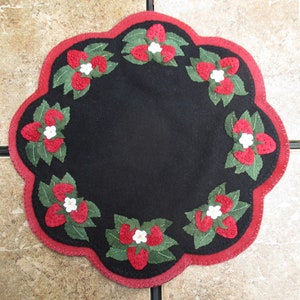 May include: A black felt round rug with a red scalloped edge. The rug is decorated with a circle of embroidered strawberries with green leaves and white blossoms.