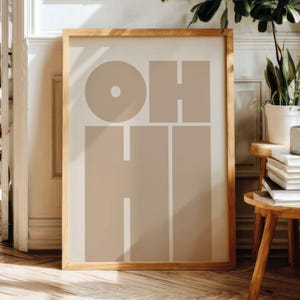 May include: A framed art print with the word "OH HI" in a minimalist design. The letters are in a beige color against a cream background, framed in a light wood. The artwork is displayed in a room with wooden floors and plants.