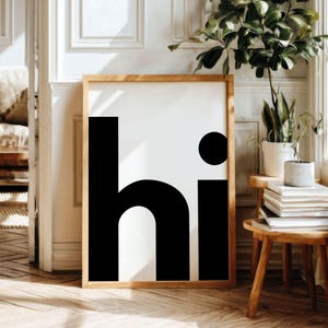 May include: A framed art print with the word "hi" in a bold, minimalist design. The black letters are set against a white background, framed by a natural wood border. The artwork is displayed in a bright, well-lit room with plants.