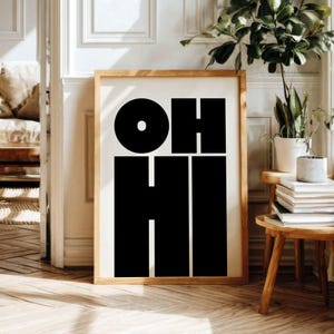 May include: A framed art print with the words "OH HI" in bold, black block letters. The artwork is displayed in a light-colored wooden frame, set against a neutral-toned wall. The minimalist design is a modern home decor piece.
