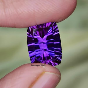Può includere: Una gemma viola rettangolare sfaccettata con un taglio a scacchiera che riflette la luce in tonalità viola intenso e porpora scuro. La pietra presenta il testo Zeenayaa World stampato in piccole lettere nere sulla sua superficie inferiore.