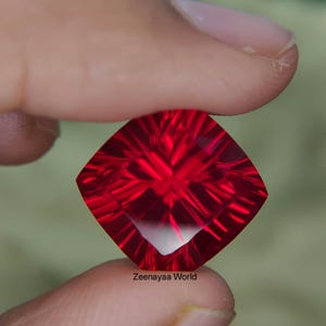 Puede incluir: Una gema de color rojo intenso y vibrante con un corte de cojín cuadrado y facetas radiantes complejas que crean un patrón de estrella. La piedra es translúcida y muy reflectante, con el texto Zeenayaa World impreso en letras pequeñas.