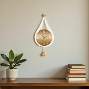 Può includere: Un orologio da parete a forma di goccia dorata e bianca con un pendolo dorato. Il quadrante dell'orologio è dorato con lancette nere e numeri bianchi. L'orologio è appeso a un cordino bianco.