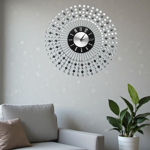 May include: A circular wall clock with a black face and silver hands. The clock is adorned with a ring of clear and black crystals. The clock is mounted on a light grey wall.