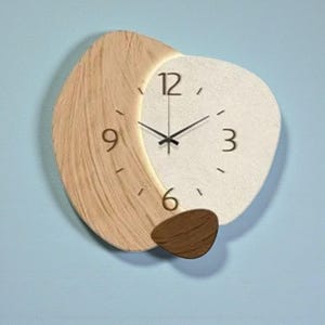 Puede incluir: Un reloj de pared moderno con un diseño único. El reloj presenta una sección de veta de madera clara a la izquierda y una esfera blanca texturizada con manecillas y números negros. Un pequeño acento de madera marrón en la parte inferior.
