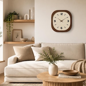 May include: A square wall clock with rounded corners, featuring a dark brown wooden frame and a cream-coloured face. The clock has black numerals at the 12, 3, 6, and 9 o'clock positions, with thin black hour and minute hands centred on the dial.