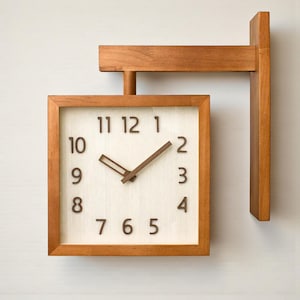 Könnte beinhalten: Eine quadratische Holzuhr mit hellem Zifferblatt und dunkelbraunen Ziffern und Zeigern. Die Uhr ist an einem Holzrahmen befestigt, der zum Aufhängen an einer Wand bestimmt ist. Die Zeiger der Uhr zeigen an, dass es ungefähr 1:10 Uhr ist.