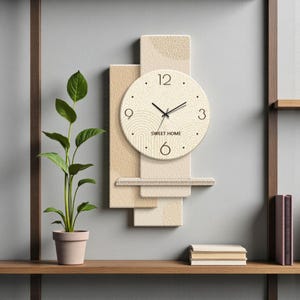 May include: A beige and tan wall clock with the text "SWEET HOME" on the clock face. The clock is mounted on a textured, layered background. A potted plant and books are on a wooden shelf below.