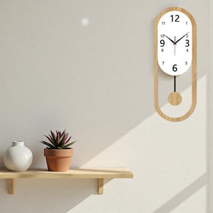 Peut inclure: Horloge murale ovale en bois clair. Le cadran blanc affiche des chiffres et des aiguilles noirs. Un pendule en bois est suspendu en dessous. L'horloge est fixée sur un mur blanc.