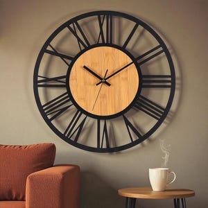 Puede incluir: Un reloj de pared grande y redondo con un marco de metal negro y números romanos. La esfera del reloj es de madera clara. El reloj está colgado en una pared de color neutro.