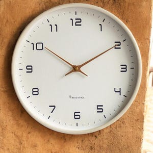 Puede incluir: Un reloj de pared blanco con números negros y manecillas marrones. El reloj tiene un diseño minimalista con el texto "MADE BY MJK" en la parte inferior. El reloj es redondo y tiene un marco blanco.