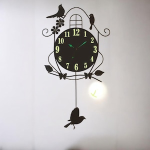May include: Black decorative wall clock with a bird and floral design. The clock face is black with glowing green numbers and hands. Two birds perch on the top, and one hangs below. The clock has a whimsical, nature-inspired theme.