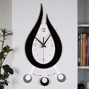 May include: A modern wall clock with a black and white teardrop design. The clock face is white with black numerals and hands. The clock has a pendulum with three circular weights. The clock is mounted on a white wall.