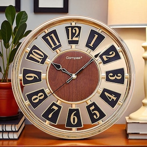 May include: A round wall clock with a gold-coloured frame and a dark wood-grain centre. The clock face features black numerals on a light background. The clock hands are black. The brand name "Compas" is printed on the clock face.