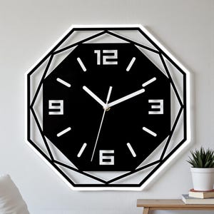 May include: A contemporary wall clock featuring a geometric design. The clock face is black with white numerals at 3, 6, 9, and 12 o'clock, and white hands. The clock is surrounded by a white geometric frame. The clock is mounted on a white wall.