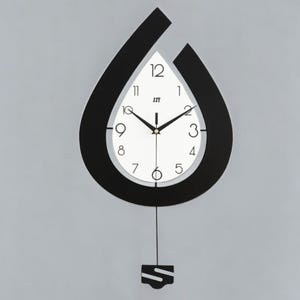 Op de afbeelding: Een moderne wandklok met een zwart, druppelvormig frame. De klok heeft een witte wijzerplaat met zwarte cijfers en wijzers. Een kleine zwarte slinger hangt eronder. De klok heeft de tekst "JST".