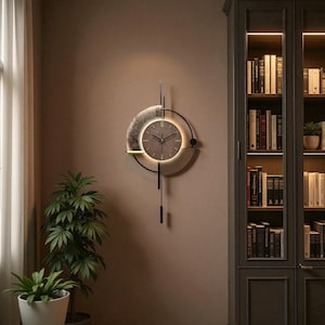 Op de afbeelding: Moderne wandklok met een cirkelvormig ontwerp. De klok heeft een grijze wijzerplaat met zwarte wijzers en een zwart metalen frame. Het heeft een decoratieve slinger en een oplichtende halo. De klok is aan een bruine muur bevestigd.