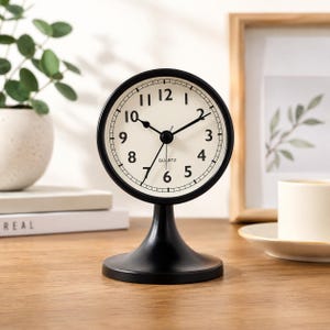 May include: A round analogue quartz table clock with a matte black frame and pedestal base. The clock face is off-white with black Arabic numerals from 1 to 12, black hour, minute, and second hands, and the word QUARTZ printed in black text.