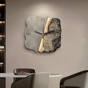 Puede incluir: Un reloj de pared único con un diseño de piedra. La esfera del reloj está dividida en dos secciones: un lado liso, gris claro y un lado texturizado, gris oscuro. Las manecillas del reloj son negras y los números 12 y 6 son visibles.