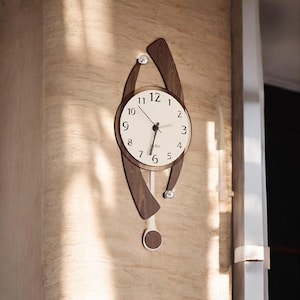 May include: A modern wall clock with a white face, black numerals, and black hands. The clock is encased in a dark brown wooden frame with a unique, curved design. A white pendulum with a circular end hangs below the clock face. Silver accents are visible.