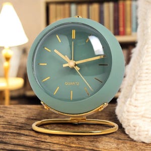 Può includere: Un orologio rotondo al quarzo verde menta con lancette dorate e base dorata. Il quadrante è visibile attraverso una copertura in vetro trasparente. La parola "QUARTZ" è stampata in basso sul quadrante. È un oggetto decorativo.