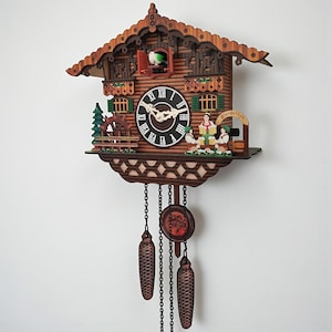 May include: A wooden cuckoo clock with a detailed, carved design. The clock features a round face with Roman numerals, a small figure in a window, and decorative elements like a water wheel and figures at a beer garden. The clock has two pinecone-shaped weights.