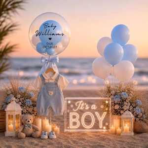 Può includere: Una scena di baby shower con una tuta blu chiaro, un orsacchiotto e palloncini. Un grande palloncino trasparente ha il testo "Baby Williams DUE OCT. 2027". Un cartello recita "It's a BOY". Candele e fiori completano l'ambientazione sulla spiaggia.