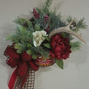 May include: A rustic floral arrangement featuring a red coffee can base, adorned with faux evergreen branches, pinecones, and berries. A large red peony and a cream-colored flower are nestled among the greenery, with a deer antler adding a natural touch. A red plaid bow completes the design.