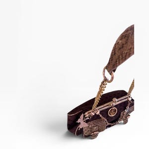 May include: A dark brown clutch bag with a gold chain strap and clasp. The bag features a butterfly charm and a small, patterned flap. The bag has a gold-coloured metal frame and a textured handle.