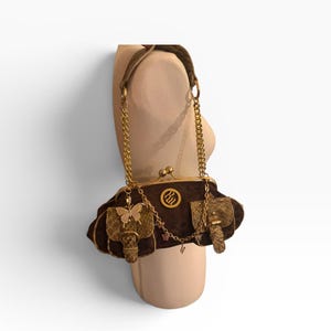 May include: A brown velvet handbag with gold chain straps and accents. The bag features gold butterfly and elephant embellishments, a gold clasp, and a decorative logo. The bag is displayed on a mannequin.
